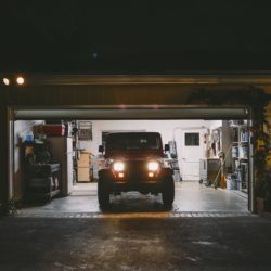 A Jeep wrangler sitting in an open garage at night with the headlights on.