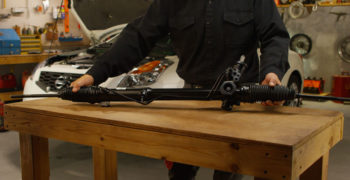 Man holding rack and pinion on a table in the garage with a Nissan Altima in the background.